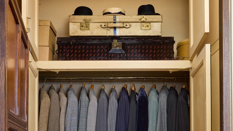 A wardrobe with a row of blue and grey suits neatly hanging up. Above them is a shelf with two trunks and three hats placed on top of it.